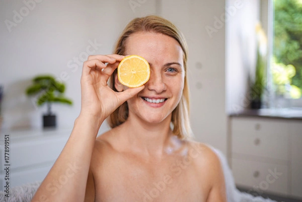 Fototapeta Smiling woman holding lemon slice in front of eye promoting healthy lifestyle