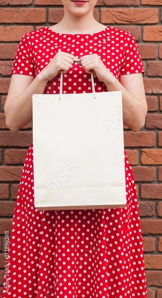Obraz Stylish woman in a retro red polka dot dress holding a blank white paper shopping bag for a mockup against a brick wall background