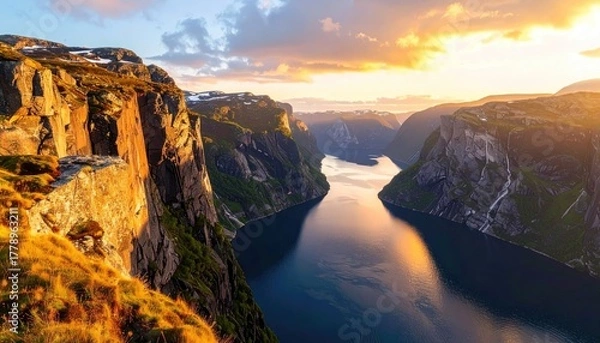 Fototapeta Dramatic golden hour sunset illuminates a winding fjord and steep cliffs with patches of snow and green moss creating a breathtaking natural landscape panorama