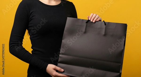 Obraz Close up of a woman in a black shirt holding a large black shopping bag against a bright yellow background
