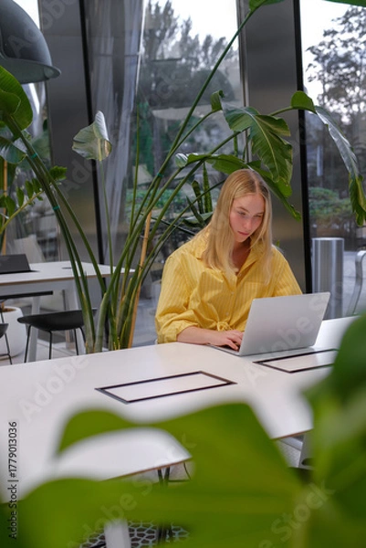 Fototapeta Gen Z freelancer with laptop working in a green coworking space among indoor plants, biophilic design; remote work, focus, productivity, flexible schedule, sustainable work lifestyle.