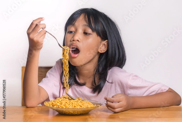 Fototapeta Hungry little girl eating noodles with a fork while sitting at table