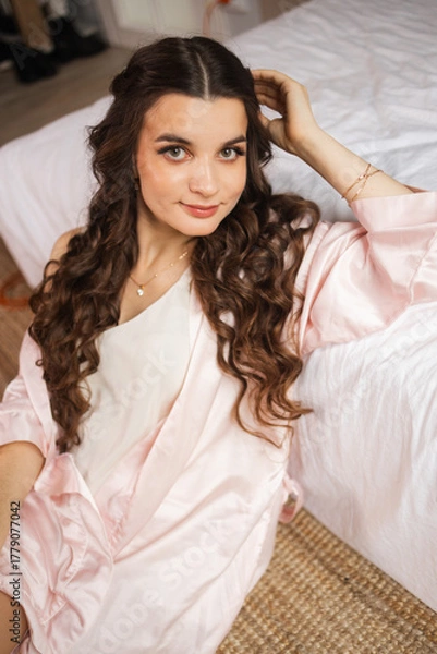 Obraz A beautiful bride poses gracefully by a sunlit window, wearing a delicate lace robe. The light highlights her serene expression and the intricate details of her bridal attire