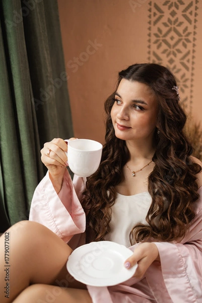 Obraz The bride is going to the wedding, drinking coffee sitting at the window. bride in a silk robe, morning of the bride, wedding day.