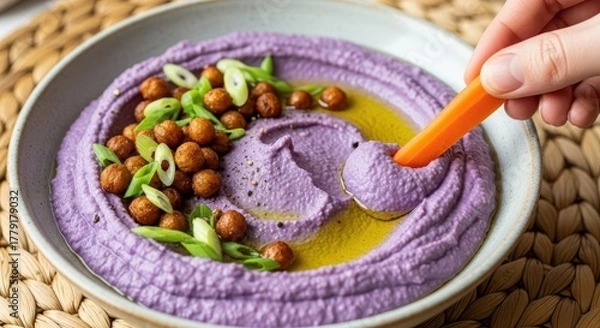 Fototapeta A hand dips a carrot stick into a bowl of vibrant purple hummus