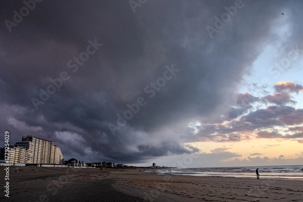 Fototapeta Nieuwpoort, West-Vlaanderen, Belgium, September 13th, 2025, A stunning beach scene with a dramatic sky during sunset, showcasing a lone figure by the tranquil shore