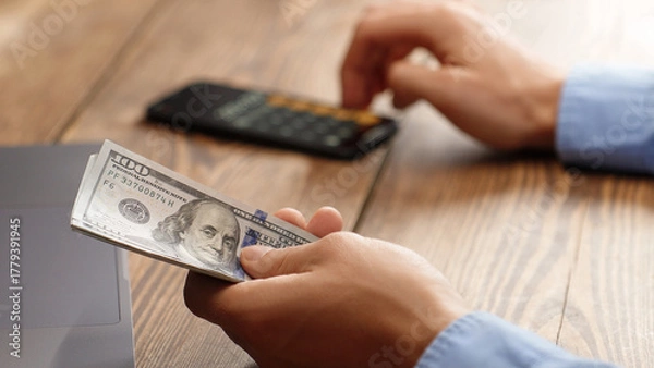 Obraz Man holding a pile of 100 dollar bills while using calculator. Financial planning, budgeting or tax calculation concept. Ideal for personal finance, business or investment content.