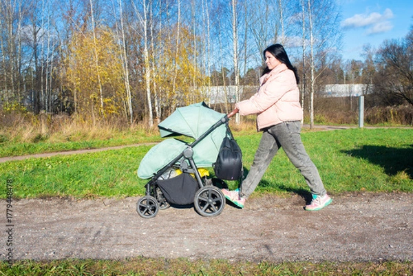 Obraz A brunette woman walks with her sleeping baby son in a green stroller in an autumn park.	