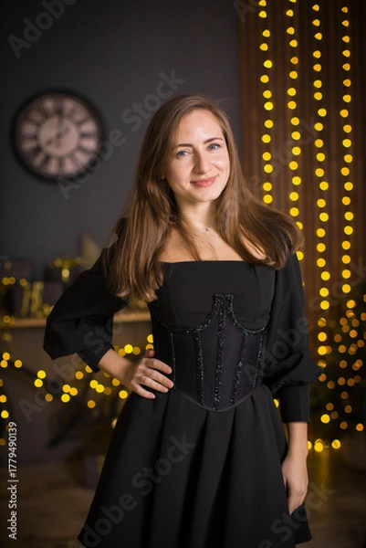 Fototapeta A woman in a black dress and stockings stands in front of a Christmas tree. She smiles and poses for the camera. The concept of holidays and Christmas.