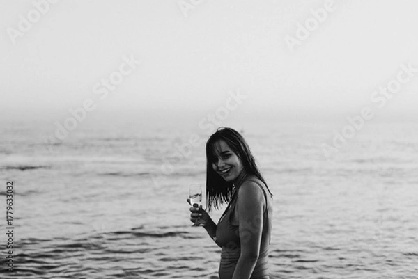 Obraz A cheerful woman in a vibrant red swimsuit stands by the seaside, holding a glass of white wine. The setting sun casts a warm golden hue over the scene, highlighting her joyous expression.