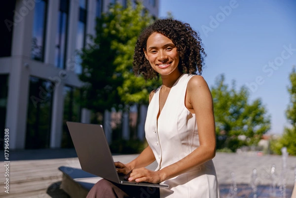 Fototapeta woman confidently working on laptop amid sunny outdoor environment with urban architecture
