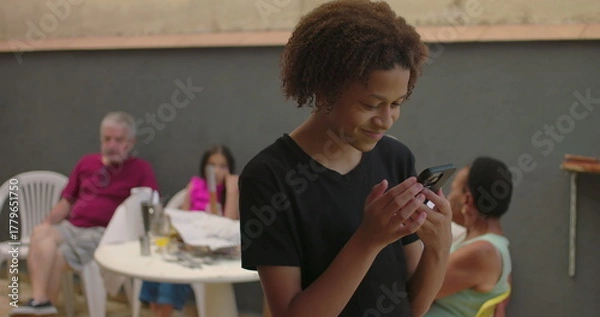 Fototapeta Teenager of African descent holding smartphone with green screen, chroma key feature displayed, smiling outdoors at a casual gathering with diverse adults in the background