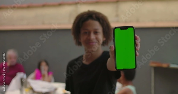 Fototapeta Teenager of African descent holding smartphone with green screen, chroma key feature displayed, smiling outdoors at a casual gathering with diverse adults in the background