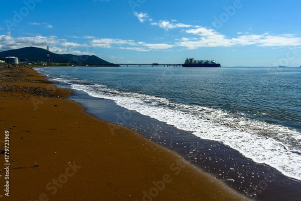 Fototapeta Sunny shoreline with gentle waves and a distant ship near hills under a clear blue sky