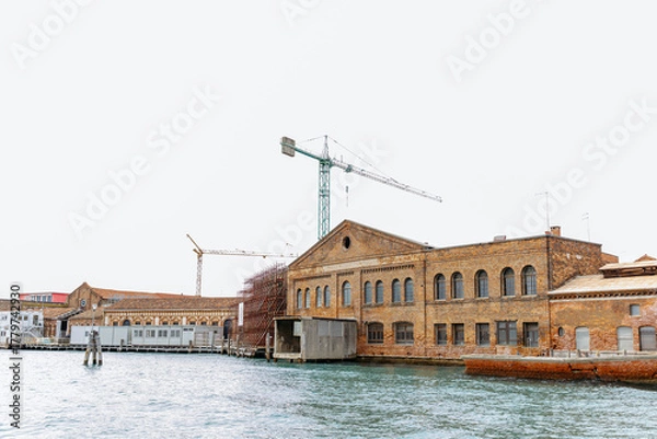 Fototapeta Murano, Italy - November 21, 2024: Scenic view of historic Murano island architecture with waterfront, showcasing traditional buildings and modern cranes, reflecting the blend of history and progress