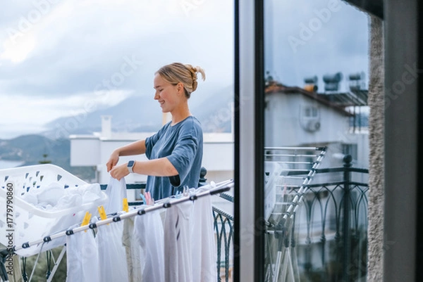 Obraz Young woman hanging laundry on balcony drying rack and clothespins, air-drying for energy saving and eco-friendly living, casual apartment lifestyle with city and mountain view, candid domestic scene