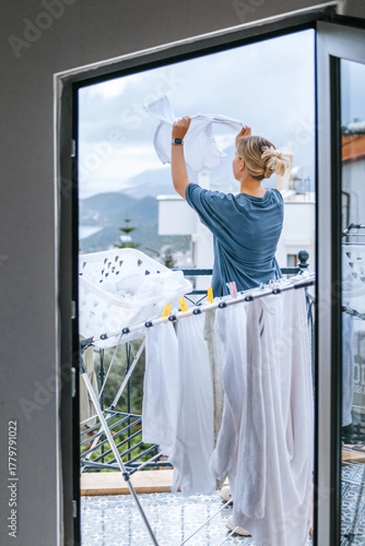 Obraz Young woman hanging laundry on balcony drying rack and clothespins, air-drying for energy saving and eco-friendly living, casual apartment lifestyle with city and mountain view, candid domestic scene