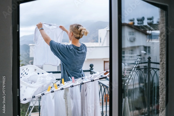 Fototapeta Young woman hanging laundry on balcony drying rack and clothespins, air-drying for energy saving and eco-friendly living, casual apartment lifestyle with city and mountain view, candid domestic scene