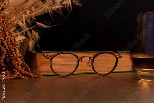 Fototapeta Stylish reading glasses resting on a vintage book, with a glass of whisky partially visible on the right. A cozy and intellectual still life scene, suggesting relaxation or evening study