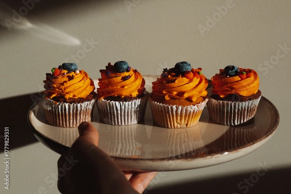 Fototapeta Delicious Halloween cupcake with orange frosting and blueberry held by a hand. A hand holds a cupcake with orange frosting, a blueberries