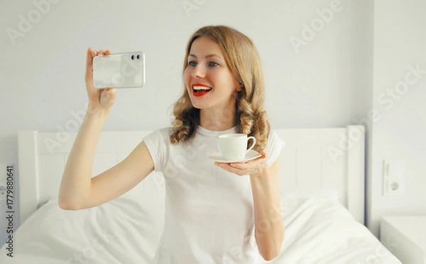 Fototapeta portrait of happy smiling young woman taking selfie with phone while lying on bed at home