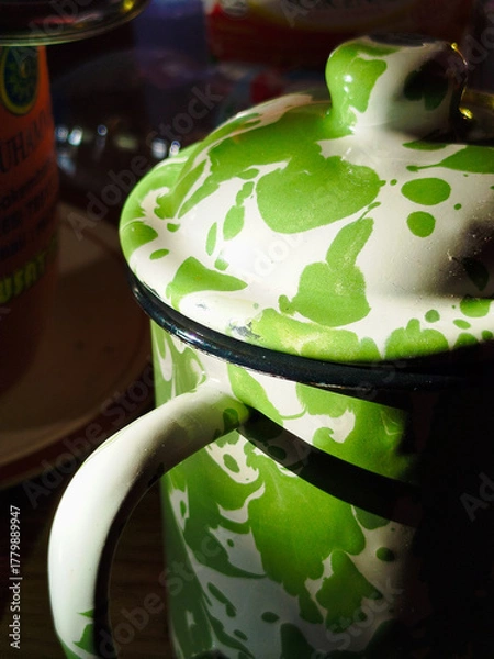 Fototapeta Closed view of a vintage-style green and white enamel teapot, featuring a splattered paint pattern and curved handle. This stylish kitchenware design is placed in a room with dramatic lighting. It's p