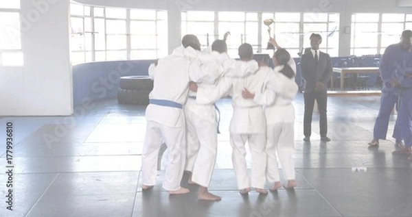 Fototapeta Cheering martial arts team gathering in training hall, lifting gold trophy and wearing white gis