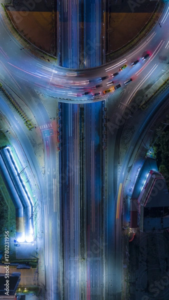 Fototapeta Top view of urban highway interchange at night with colorful light trails from fast-moving cars. Modern city traffic captured by drone in long exposure, showing speed, energy, and infrastructure 