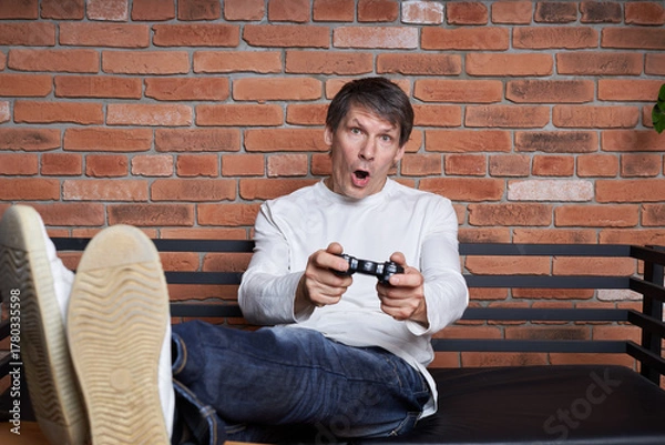 Obraz A middle-aged man gamer holds a gamepad and plays an online game on a computer.