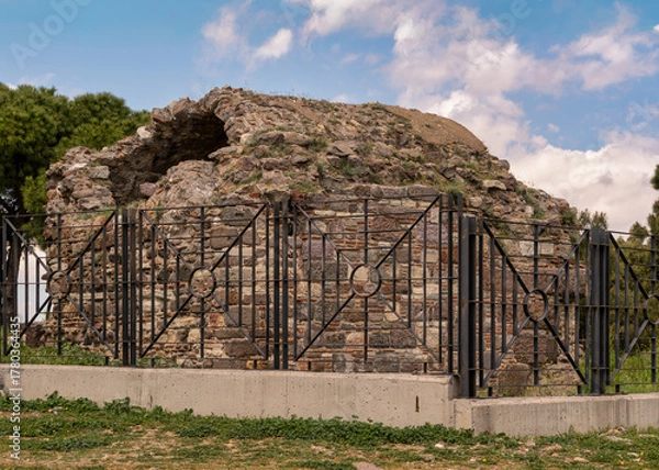Fototapeta Ruins of Castle Masjid, in Kadifekale Castle, Izmir, Turkey. Ancient stone mosque remains, partially overgrown, behind a protective fence under a cloudy sky.