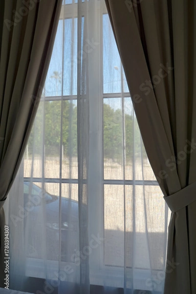 Fototapeta A softly lit window framed by beige curtains with tiebacks and sheer white drapes, offering a view of greenery through multi-pane glass.