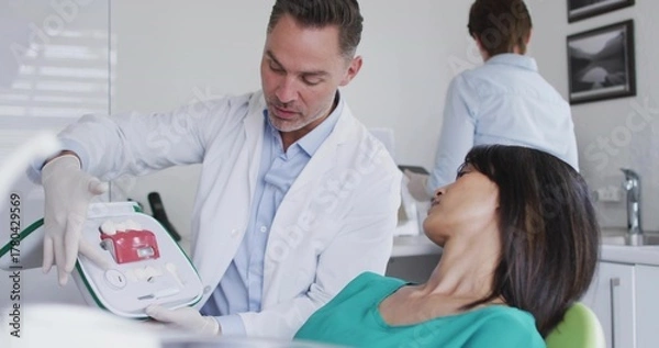 Obraz Dentist in white coat holding dental model and explaining to patient in clinic, with dental chair