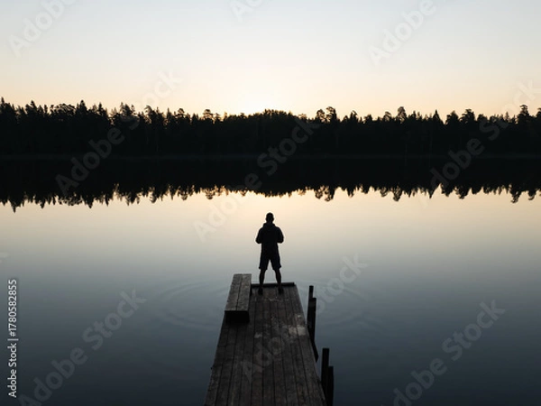 Fototapeta Aerial drone view of a person's silhouette on a wooden pier overlooking a tranquil Estonian lake with perfect forest reflections at sunset.