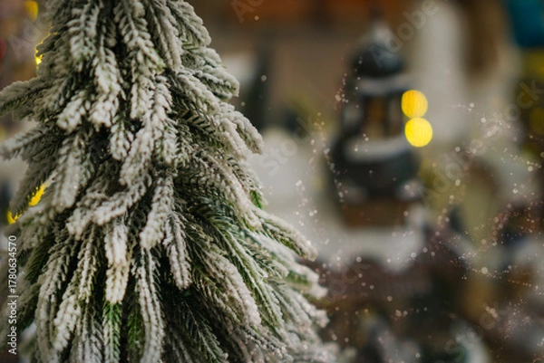 Fototapeta a snow covered miniature pine tree with bokeh lights and soft snowfall effect in a Christmas village concept of holiday advertising, winter celebration, festive greeting