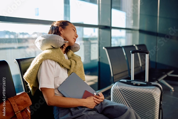 Obraz Tired woman resting comfortably with a neck pillow and luggage in an airport departure lounge, waiting for her flight