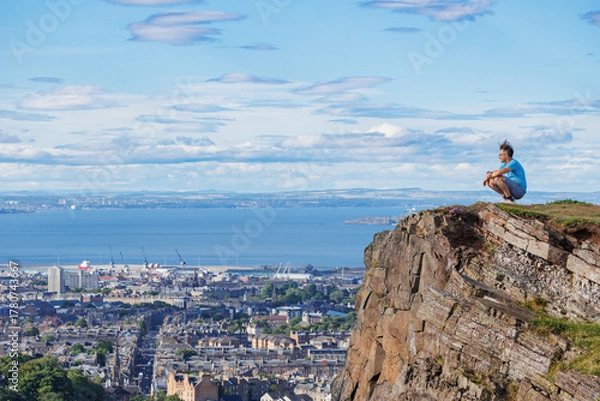 Fototapeta Tourist perched on cliff, taking in urban landscape and ocean