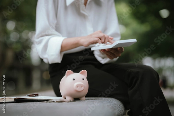 Obraz A person saves money by putting coins into a pink piggy bank while holding a calculator, symbolizing financial planning, saving, and budgeting.