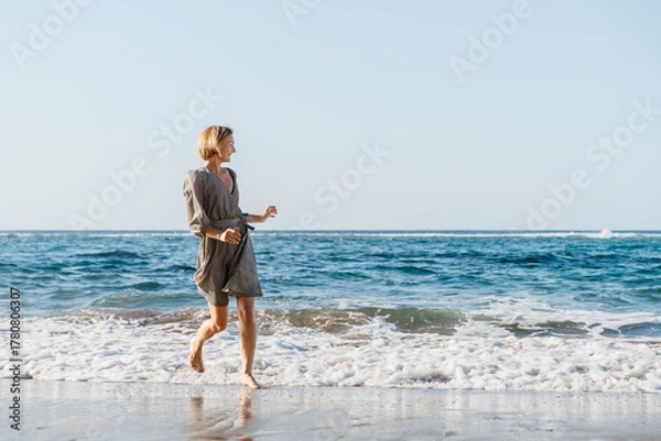 Fototapeta Happy woman runs and jumps by the ocean. Concept freedom and carefree vacation. Beach Holidays. Carefree woman splashing water having fun on the beach. Young girl enjoying fresh air on ocean shore.