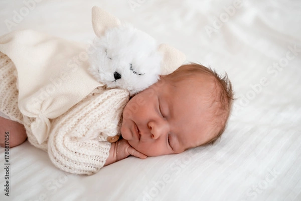Fototapeta newborn baby sleeping on a white bed at home with a soft toy, close-up portrait of a newborn baby sleeping in a white knitted bodysuit, space and room for text