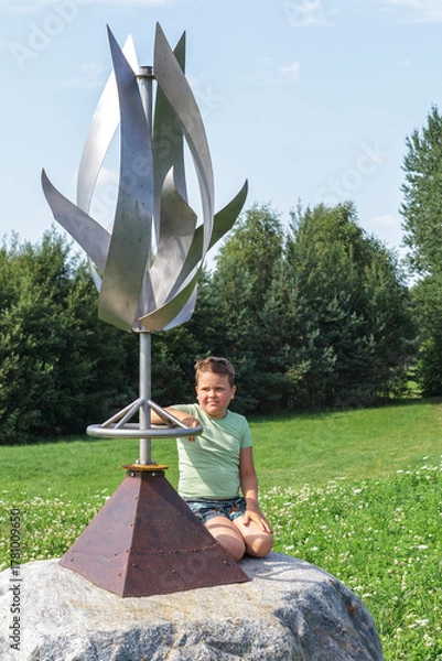 Fototapeta A little boy sits on a stone next to the wind sculpture and looks at us