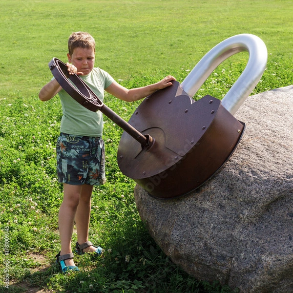 Fototapeta A little boy on the playground is trying hard to unlock a giant lock with a large metal key. Square photo