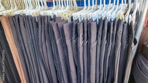 Obraz a clothing rack with several pairs of pants hanging on white plastic hangers. Most of the pants are dark-colored—mainly black or dark blue.The pants are neatly arranged and folded over the hangers.