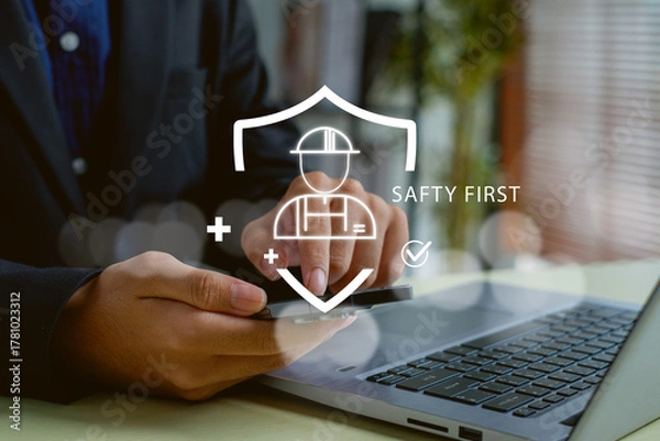 Fototapeta professional using a smartphone and laptop to manage workplace safety digitally. Virtual safety first icon with helmet and shield representing protection, risk control, and smart safety management.