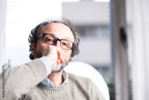 Fototapeta Portrait of a thoughtful middle-aged man with glasses sitting indoors by a window. Casual sweater, soft light, introspective mood. Concept of reflection, thinking or modern lifestyle.