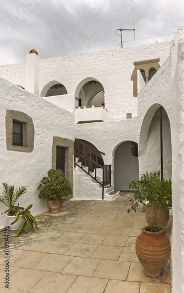 Fototapeta Stone courtyard, whitewashed walls and arched doorways,  cobblestone flooring, potted plants,  small tree and flowers, in the Monastery of Saint John the Theologian on the island of Patmos, Greece.
