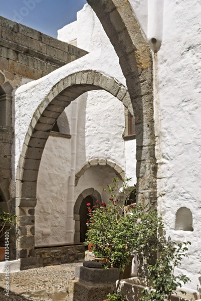 Fototapeta Stone courtyard, whitewashed walls and arched doorways,  cobblestone flooring, potted plants,  small tree and flowers, in the Monastery of Saint John the Theologian on the island of Patmos, Greece.
