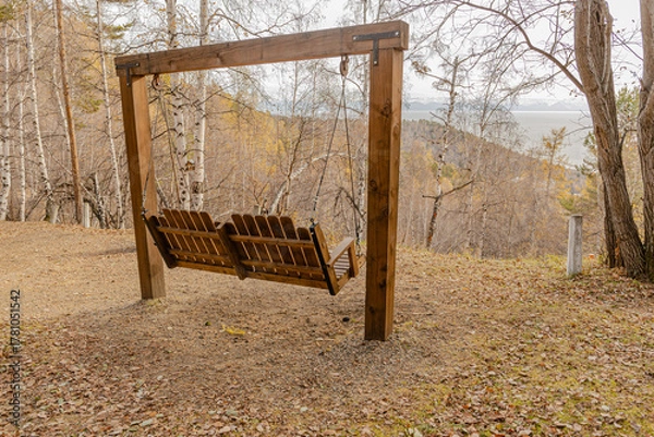 Fototapeta Wooden bench swing hangs from a sturdy frame in a quiet autumn park overlook distant hills and a hazy valley and a river or lake beyond the trees.