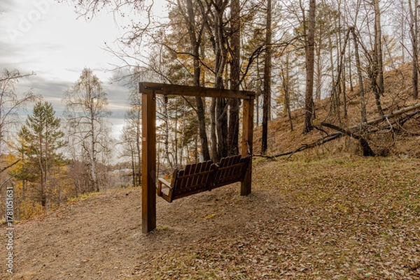 Fototapeta A wooden bench swing hangs from a sturdy frame in a sunlit autumn forest on sloped hill overlook autumn trees and a river or lake.  National Park at Baikal Lake