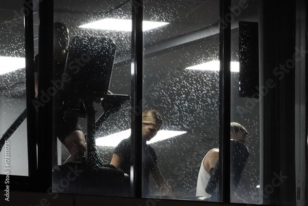 Fototapeta Young people doing strength training in a gym. A view from the street through the window of a gym with fitness enthusiasts