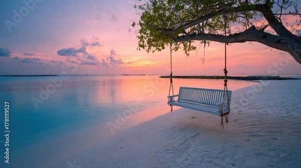 Fototapeta A rustic rope swing hanging from a tree branch on a sandy beach during a vibrant orange and pink sunset over calm water.